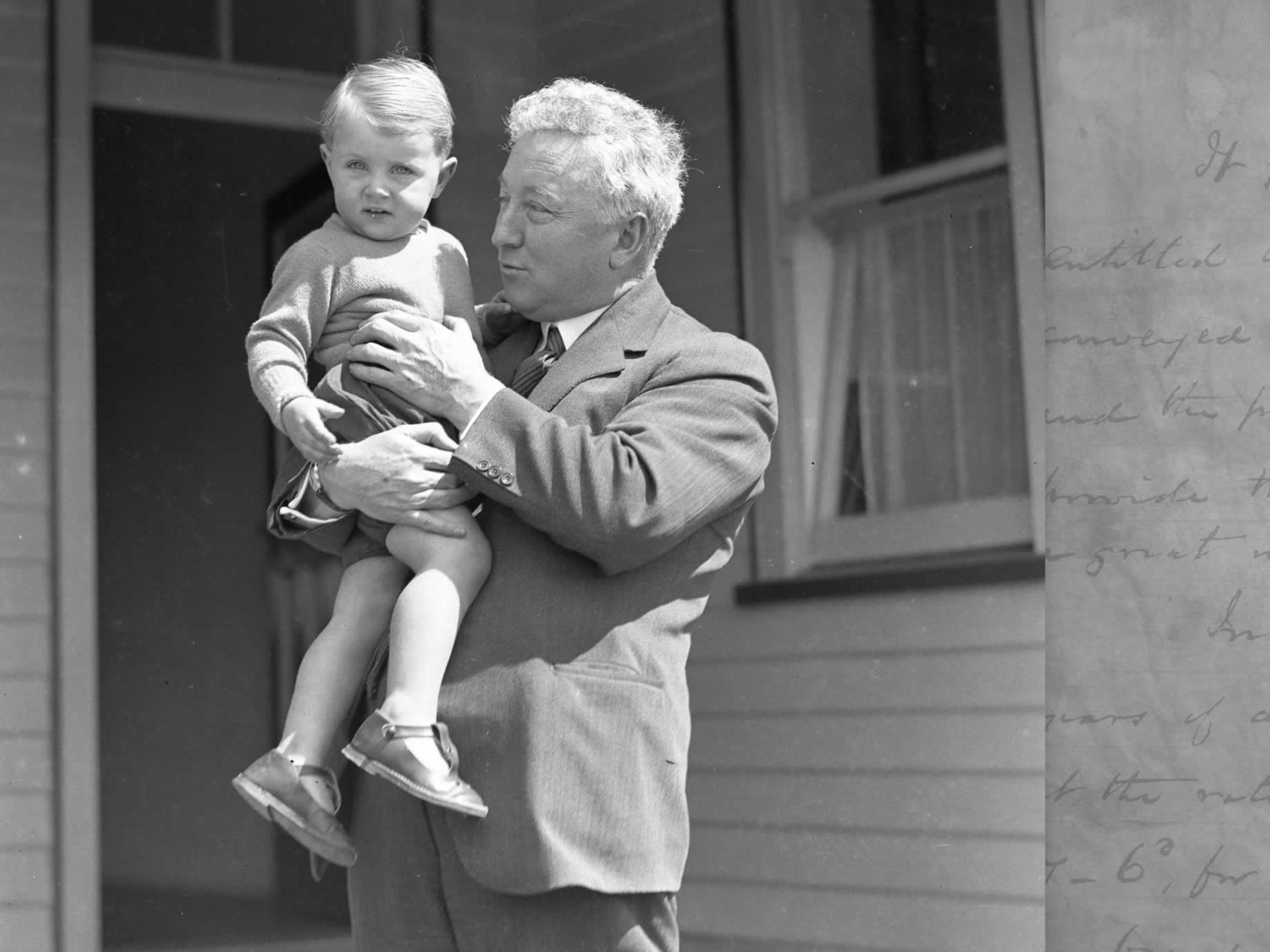 Joe Lyons devant la porte arrière de sa maison avec un jeune enfant au milieu des années 1920